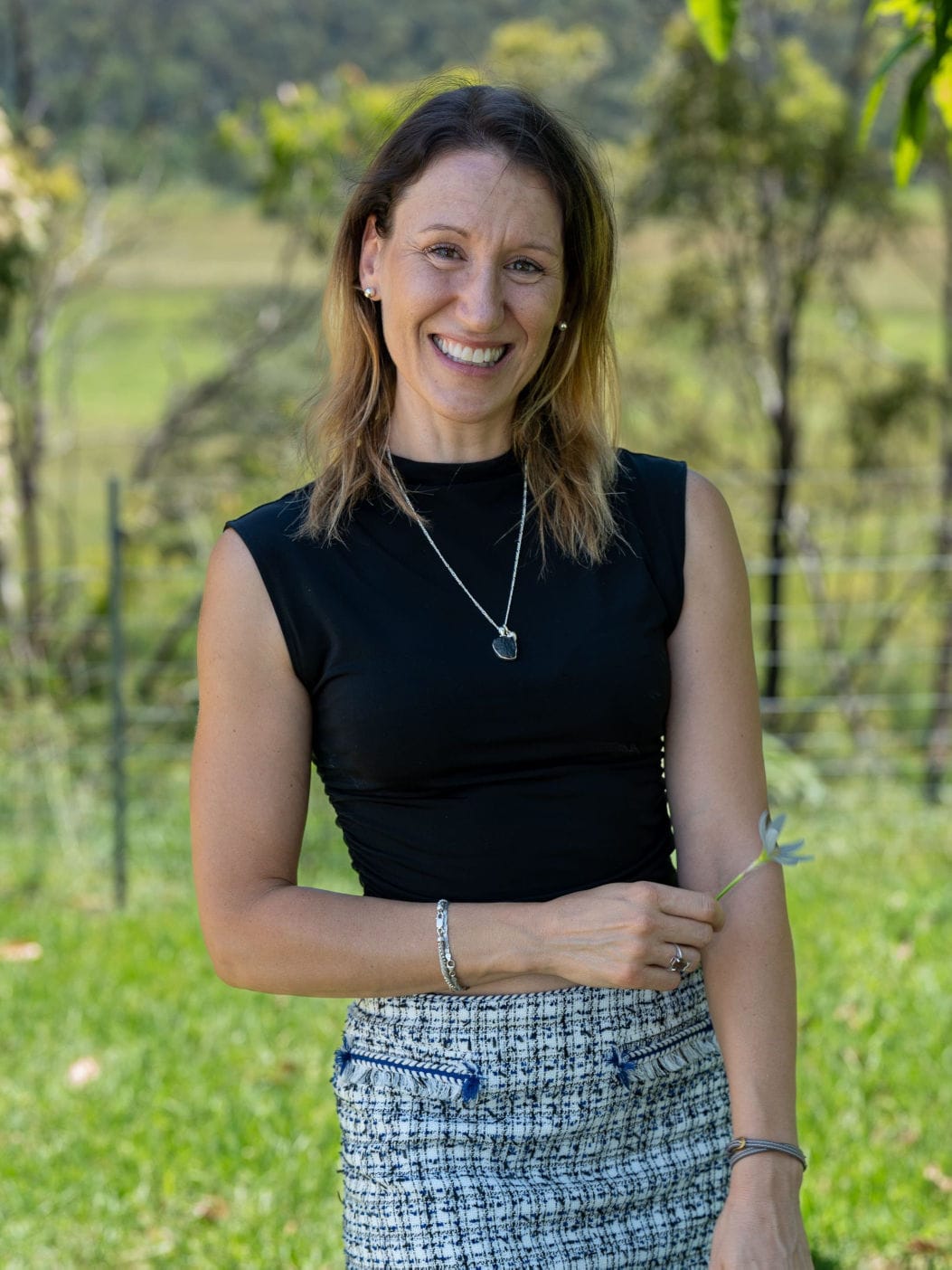 a potrait photograph of Laure Mignot, owner of nrg healing. She has, shoulder length, dark brown hair with accents of blonde and is wearing a black fitted top and a white and blue patterned skirt. She is holding a flower in her right hand.
